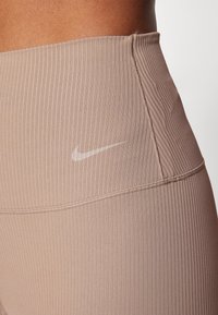 Leggings côtelés marron clair avec une taille haute et un logo Nike blanc contrastant. Des détails de couture visibles mettent en valeur la texture.