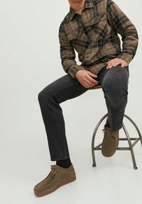 Man med brun rutig skjorta, svarta jeans och bruna mockaskor som sitter på en metallpall mot en neutral bakgrund.