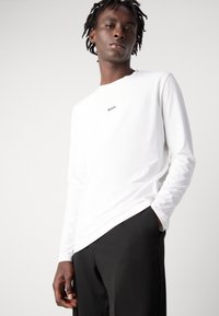 Camisa branca de manga longa com gola redonda, apresentando um pequeno logótipo "BOSS" no peito. Combinada com calças pretas. Textura de tecido suave.