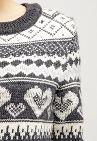 Neulottu villapaita tummanharmaana ja valkoisena, pyöreällä kaula-aukolla. Koristeena geometrisia ja sydänkuvioita, jotka esittelevät teksturoitua, kodikasta muotoilua.