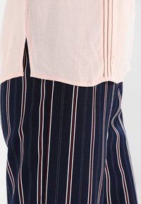 Light pink textured top with a rounded hem, paired with navy blue trousers featuring vertical stripes in red and white, showing a relaxed fit.