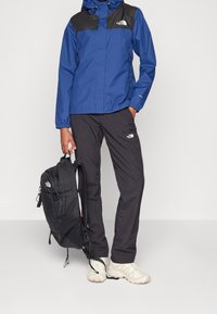 Veste imperméable bleue avec des accents noirs, pantalons noirs et un sac à dos noir. Les chaussures sont beiges avec une semelle texturée. Design minimaliste.