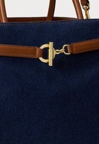 Granatowa torba o gładkim materiale, z brązowymi skórzanymi uchwytami i złotym okrągłym zapięciem. Dodatki obejmują kontrastowe szwy.