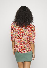Blouse à fleurs avec des fleurs rouges, orange et jaunes sur un fond clair ; manches bouffantes et coupe décontractée ; associée à une jupe verte.