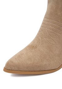 Beżowy zamszowy botek za kostkę z ostrym noskiem i subtelnym haftem w kształcie liścia. Gładka tekstura i niska, brązowa gumowa podeszwa.