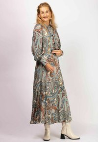 Long, patterned dress with paisley design in blue, brown, and white. Features long sleeves, a tie at the neckline, and a flowy silhouette. Paired with beige ankle boots.