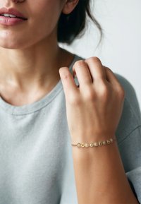 Goldarmband mit abwechselnd glatten und strukturierten Perlen sowie einem zarten Design, getragen am Handgelenk vor einem hellgrauen Hintergrund.