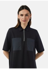 Zwart polo shirt met korte mouwen, voorzien van een rits aan de voorkant en twee gestructureerde zakken op de borst. Zachte stof met een matte afwerking.