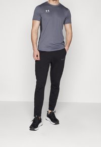Grå idrottströja med korta ärmar och logotyp på bröstet, tillsammans med svarta sweatpants med en diskret logotyp och svarta sneakers.
