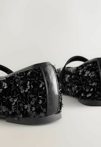 Zapatos negros con acabado de charol, adornados con lentejuelas negras y acentos texturizados, con una punta redondeada y un tacón bajo.