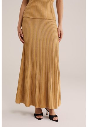 Nő, aranyszínű bordázott kötött maxi szoknyát visel, fekete nyitott orrú magassarkú cipővel párosítva, egy egyszínű bézs háttér előtt áll.