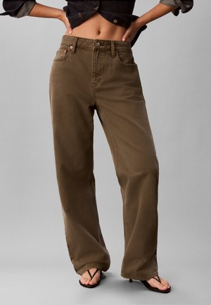 Persoon draagt losse bruine broek met hoge taille en zwarte teenslippers, staand tegen een effen grijze achtergrond.