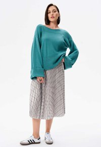 Turquoise gebreide trui met wijde mouwen, gecombineerd met een geplooide, gebloemde beige rok. Witte sportschoenen met zwarte strepen maken de outfit compleet.