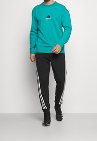 Sweat-shirt turquoise avec logo Adidas noir, associé à un pantalon noir fuselé avec des bandes blanches sur les côtés et des chaussures de sport noires.