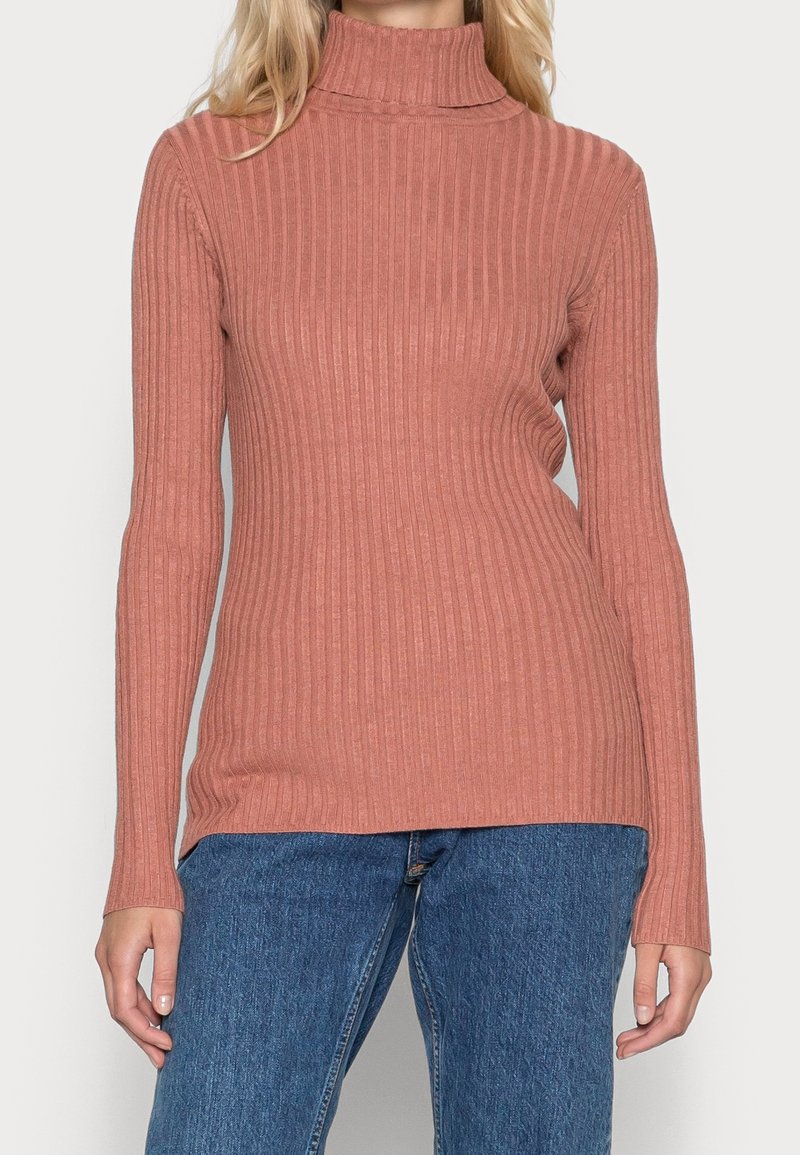 Nainen pukeutunut ribattyyn, pitkähihaiseen, haalean vaaleanpunaisiin korkeakauluksiseen villapaitaan, joka on yhdistetty sinisiin farkkuihin.