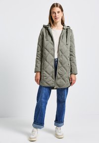 Veste matelassée olive avec capuche, fermeture éclair frontale, poches latérales et cordons de tirage réglables, associée à un jean bleu retroussé et des baskets blanches.