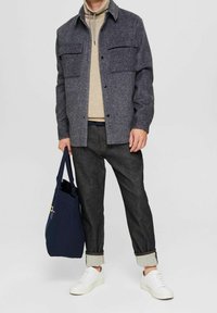 Veste en laine grise mélangée sur un pull en tricot beige, jeans foncés, baskets blanches, et un sac fourre-tout marine avec une touche colorée.