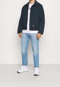 Chaqueta recortada azul marina con cuello puntiagudo, tela tejida y cremallera frontal; combinada con una camiseta blanca y jeans azul claro. Zapatillas blancas.