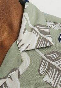 Camisa de manga corta de tela verde, con un estampado floral en beige y azul, con cuello puntiagudo y textura suave.