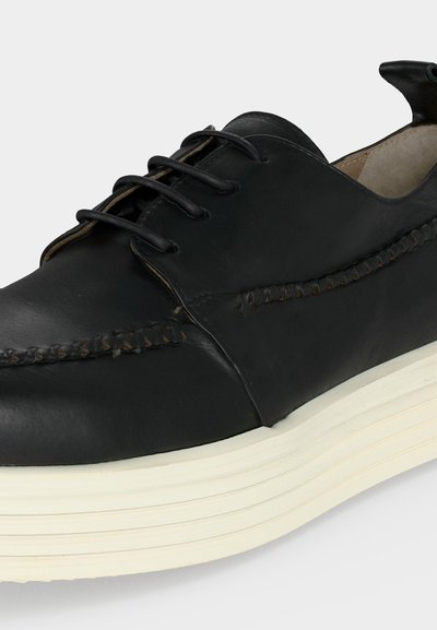 Μαύρο δερμάτινο παπούτσι με κορδόνια, χοντρή λευκή ριγωτή σόλα και διακοσμητική ραφή κατά μήκος των πλευρών και της περιοχής των δαχτύλων.