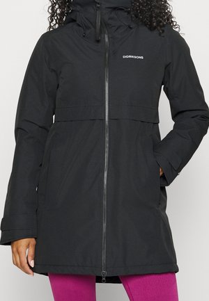 Femme portant un imperméable à capuche noir Didriksons avec fermeture éclair frontale et poches, associé à des leggings côtelés rose vif.