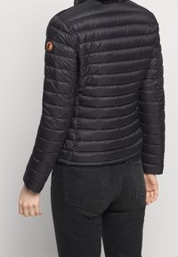 Une personne est vue de dos, portant une veste rembourrée noire avec un logo sur la manche, associée à un jean foncé. L'arrière-plan est neutre.
