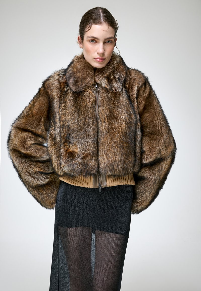 Femme portant une veste en fausse fourrure marron avec une fermeture éclair, associée à une jupe noire transparente, debout devant un fond clair uni.