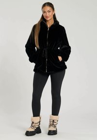Giacca nera in finta pelliccia con zip e vita regolabile, dotata di colletto alto e tasche frontali, abbinata a leggings neri e stivaletti soffici.