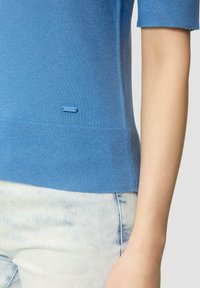 Vue de côté d'une personne portant un pull bleu à manches courtes et un jean délavé, se concentrant sur l'ourlet du pull et une petite étiquette de marque.