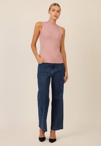 Ärmelloser, rosa Strickoberteil mit hochgeschlossenem Kragen, kombiniert mit blauen,weiten Jeans mit dezentem Detail und abgerundet mit schwarzen spitzen Absätzen.