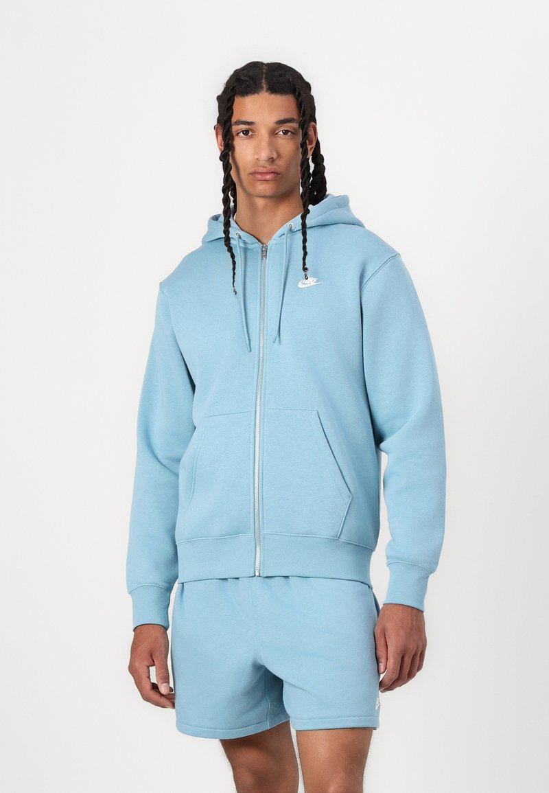 Lyseblå zip-up hettegenser med hette og frontlommer, laget av mykt stoff. Kombinert med matchende shorts, begge med en liten logo.