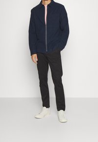 Marinblå zip-jacka med krage och en ficka på bröstet, tillsammans med svarta byxor och vita sneakers. Slät tygstruktur.