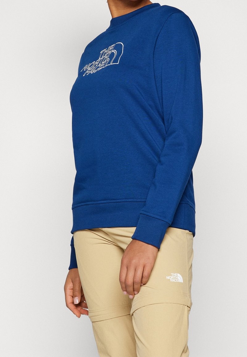 Personne portant un sweat-shirt bleu et un pantalon beige, tous deux avec le logo The North Face, debout avec une main détendue le long du corps.