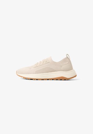 Sneaker sportiva di colore beige chiaro, lavorata a maglia, con chiusura frontale a lacci, suola ammortizzata e suola esterna in gomma naturale, mostrata di profilo su sfondo bianco.