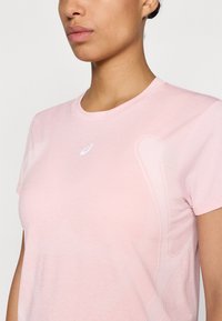 Lys rosa kortermet T-skjorte laget av myk stoff, med rund hals og subtil tekstur, samt en hvit logo på brystet.