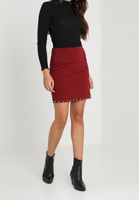Rode aansluitende rok met parel-details aan de onderrand, gecombineerd met een zwart top met lange mouwen en zwarte enkellaarsjes. Glad textuur van de stof.