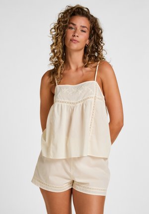 Frau mit lockigem Haar, die ein cremefarbenes besticktes ärmelloses Oberteil und passende Shorts trägt, steht vor einem schlichten Hintergrund.
