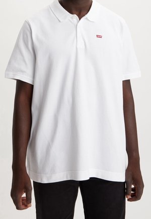 Donkerhuidig persoon draagt een wit Levi's kortermig poloshirt met kraag en rood logo op de linkerborst, gecombineerd met een zwarte broek.