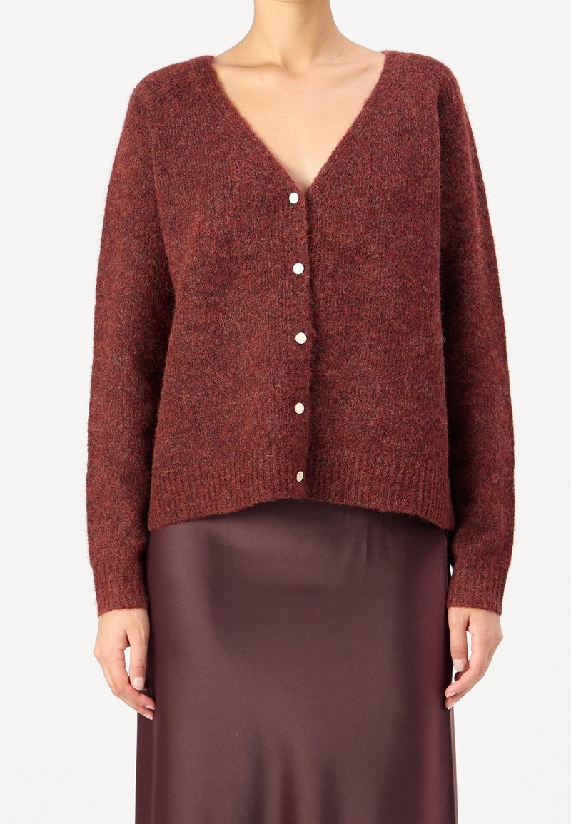 Femme portant un cardigan bordeaux en fausse fourrure, boutonné avec un col en V et des manches longues, sur une jupe en satin assortie, montrée du cou jusqu'à mi-cuisse.