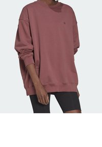 Person som bär en oversized sweatshirt i dammrosé med långa ärmar och svarta åtsittande shorts, med handen avslappnat vid sidan mot en enfärgad bakgrund.