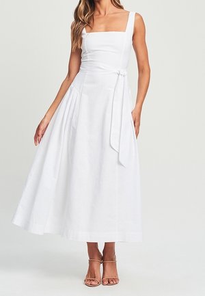 Vrouw draagt een mouwloze witte midi-jurk met een vierkante halslijn, een zijstrikdetail en beige sandalen met bandjes.