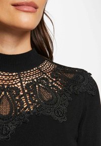Top negro de cuello alto con intrincados detalles de encaje alrededor del cuello, con tejido acanalado texturizado y una silueta ajustada.