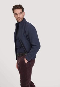 Marineblauwe zip-up trui met hoge kraag en geribde manchetten, gedragen over een zwarte shirt en gecombineerd met bordeauxrode corduroypantalon.