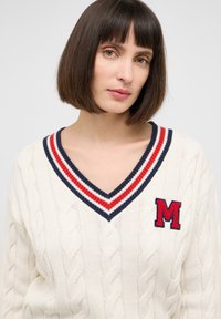 Kremowy sweter dzianinowy z dekoltem w kształcie V, z czerwonym i granatowym paskiem oraz czerwonym haftowanym logo "M" na lewej piersi.