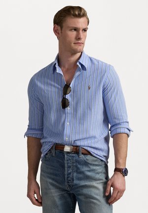 Lichtblauw overhemdd met witte verticale strepen, opgerolde mouwen en een logo op de borst. Gecombineerd met denim jeans en accessoires zoals een zonnebril.