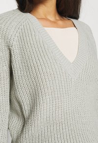 Jasnoszary sweter dzianinowy z teksturowanym wzorem, dekoltem w kształcie litery V oraz prążkowanymi detalami na dekolcie i rękawach.