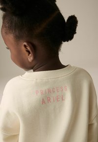 Krémový svetr s potiskem "PRINCESS ARIEL" v růžové barvě na zadní straně. Měkká tkanina s kulatým výstřihem a žebrovanými manžetami.