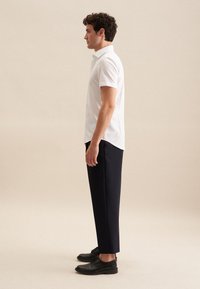 Witte polo met korte mouwen, gecombineerd met donkerblauwe broek en zwarte schoenen. Gladde stof en op maat gemaakte pasvorm. Zijaanzicht.