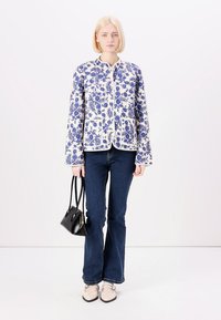 Floral-patterned quilted jacket in white and blue, featuring a round neckline and front pockets, paired with dark denim flared jeans and beige shoes.