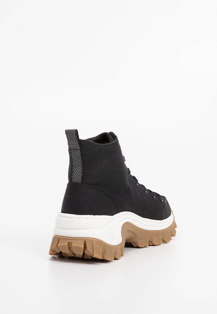 Schwarzer High-Top Sneaker mit dicker weißer Zwischensohle und dicker brauner Profilsohle, von hinten links auf einem weißen Hintergrund betrachtet.